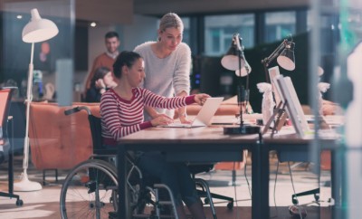 Disabled businesswoman in a wheelchair working in a creative office. Business team in modern coworking office space. Colleagues working in the background at late night. Inclusion and handicap concept.