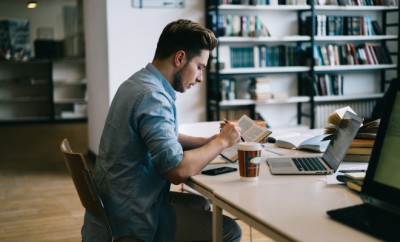 Pensive male student sitting at table in college library reading book for education and knowledge, clever hipster guy spending time on autodidact using dictionary and laptop for language courses