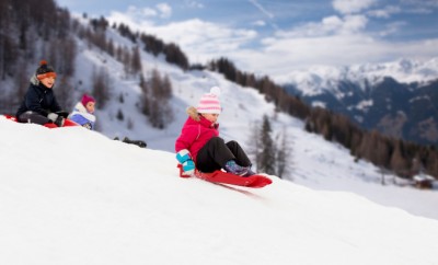childhood, sledging and season concept - group of happy little kids sliding on sleds down snow hill in winter over alps mountains on background