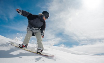 Young female snowboarder running down the mountain slope on the background of mountain and clear blue sky