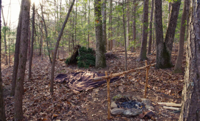Primitive Bushcraft survival debris hut with campfire ring outside. Blanket, shelter, fire in the forest.