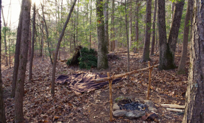 Primitive Bushcraft survival debris hut with campfire ring outside. Blanket, shelter, fire in the forest.